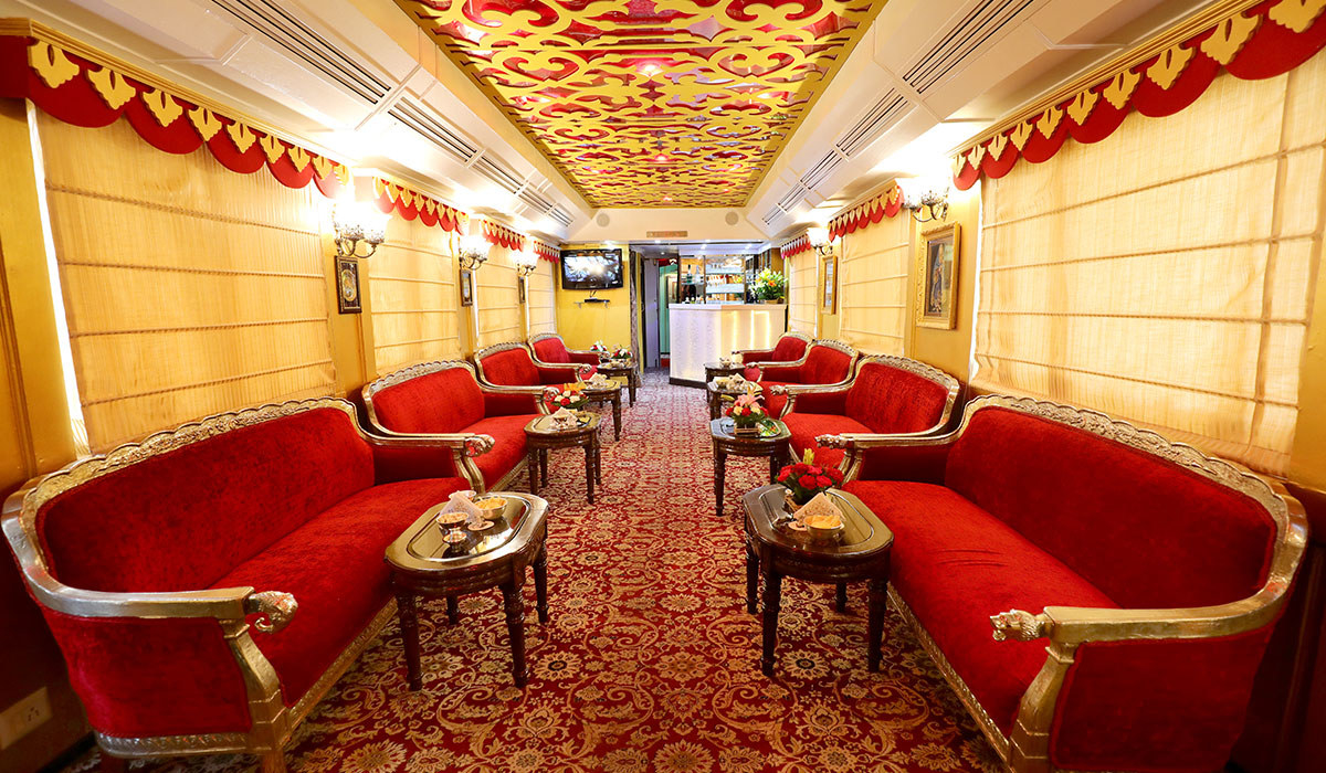 A luxurious train carriage with an ornate ceiling, plush red and gold couches, tables with flowers, glowing lamps, and a patterned carpet