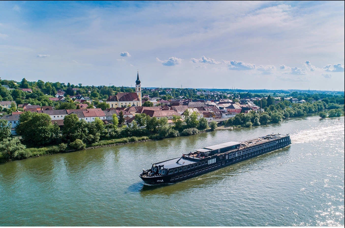 A long boat travelling down the river, on the banks is a small European town