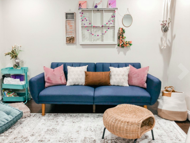 Reviewer&#x27;s navy blue velvet futon adorned with pink, white, and rust-tone accent pillows in a room filled with wicker tables and baskets
