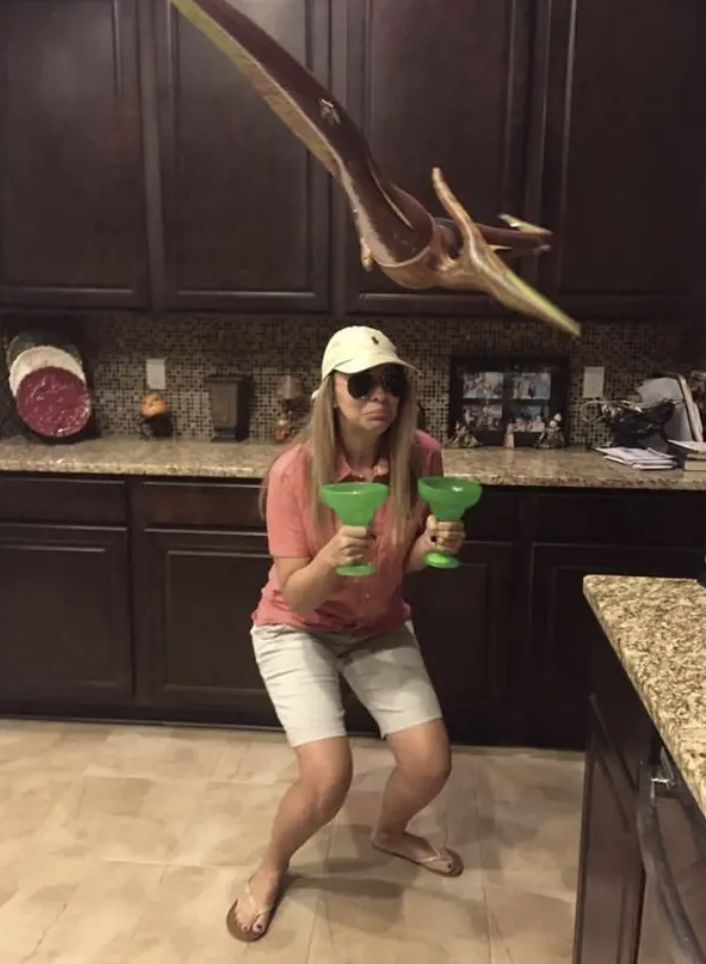 Someone dressed in summer clothes, holding two margarita glasses, with a fake dinosaur flying over head