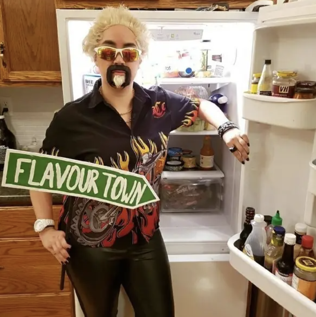 Someone dressed as Guy Fieri (goatee, dyed hair, flames t-shirt), standing next to an open fridge
