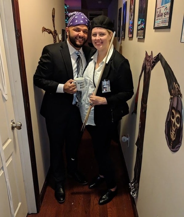 Two people standing next to each other in suits. One is wearing a bandana on their head and holding a "World's Best Boss" mug, and the other is wearing a newsboy hat and holding a players cue.