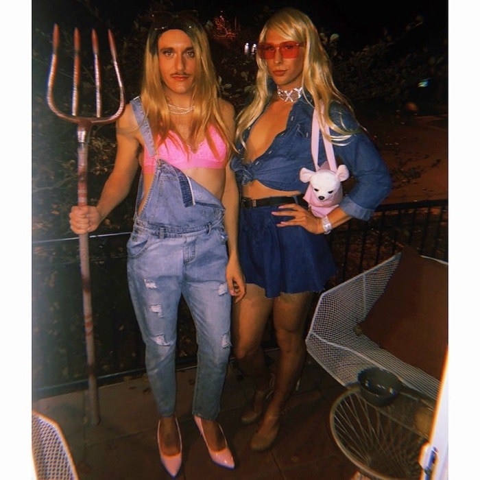 Two men dressed in overalls, heels, and blonde wigs. One is holding a pitchfork and the other is holding a fake puppy. 
