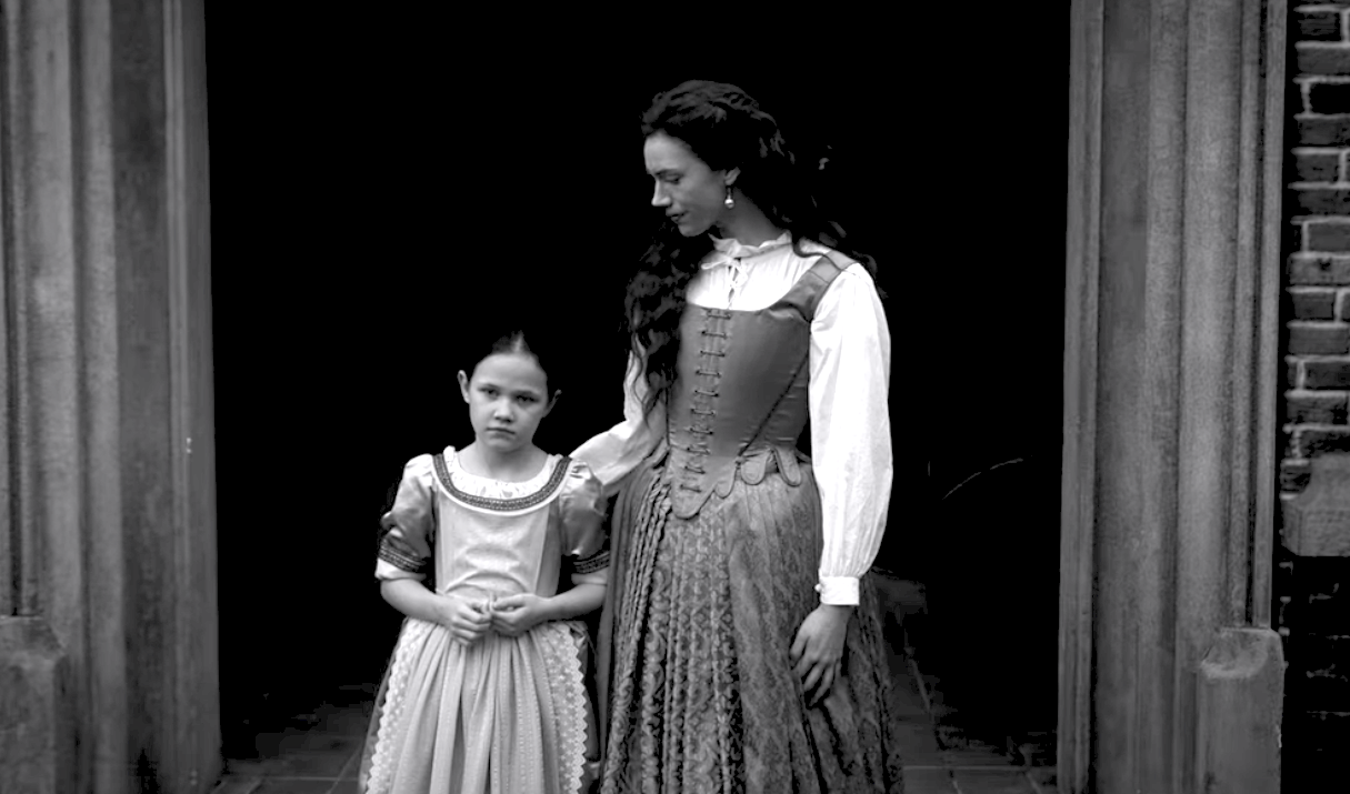 A black and white image of a woman and a young girl standing in front of a front door in old timey clothes