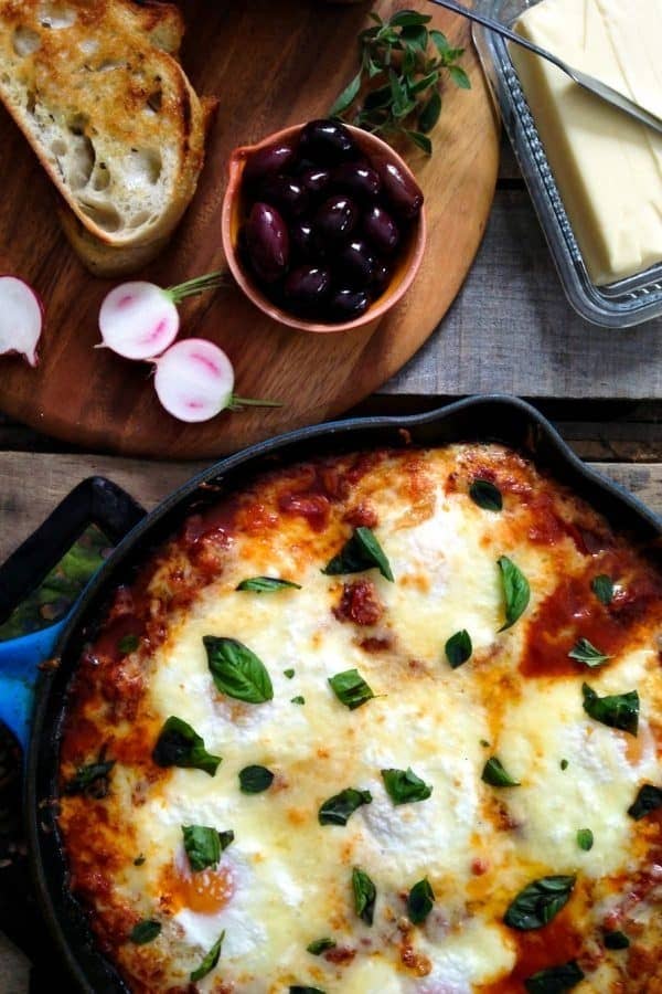 A cast iron skillet filled with cheesy baked eggs with sliced bread and olives on the side.