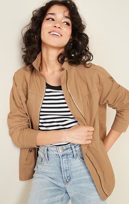 Model wearing the jacket in crumb it on down light brown color over a striped shirt tucked into light blue jeans