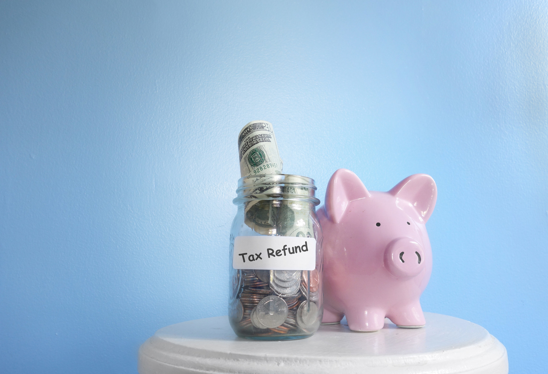 A stool holds a pink ceramic piggy bank and a clear jar filled with money and a sticker that reads &quot;tax refund&quot;