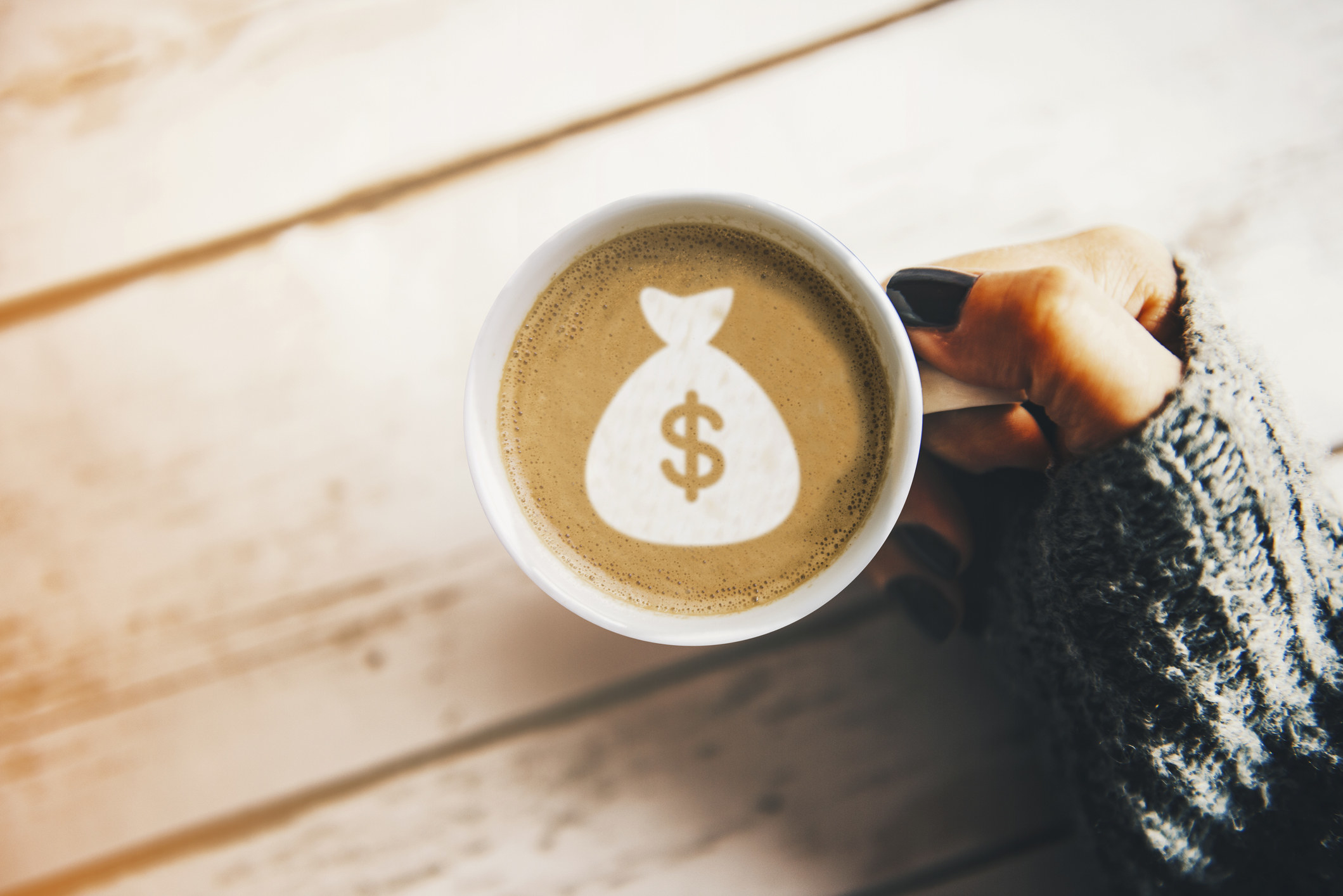 A hand holds a cup of coffee with a foam design of a money bag