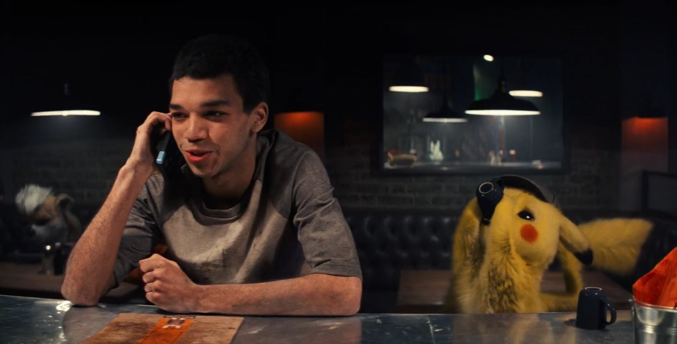 A man on the phone in a diner, next to a yellow Pikachu drinking coffee