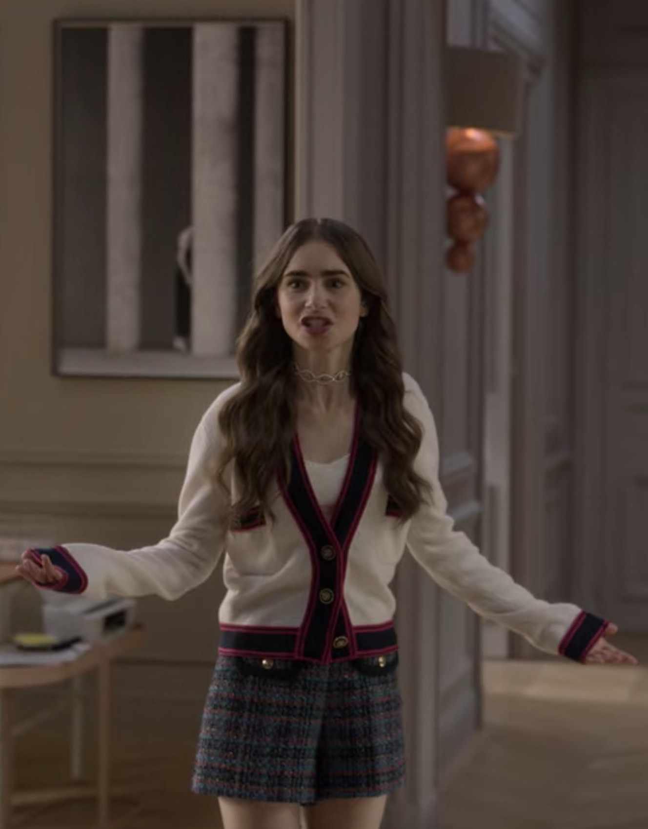 Emily gestures angrily while wearing an oversized red and cream cardigan and plaid shorts