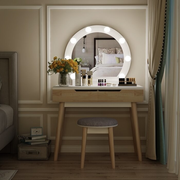 wood vanity with hollywood-style LED bulbs around a round mirror. A gray stool sits below the vanity