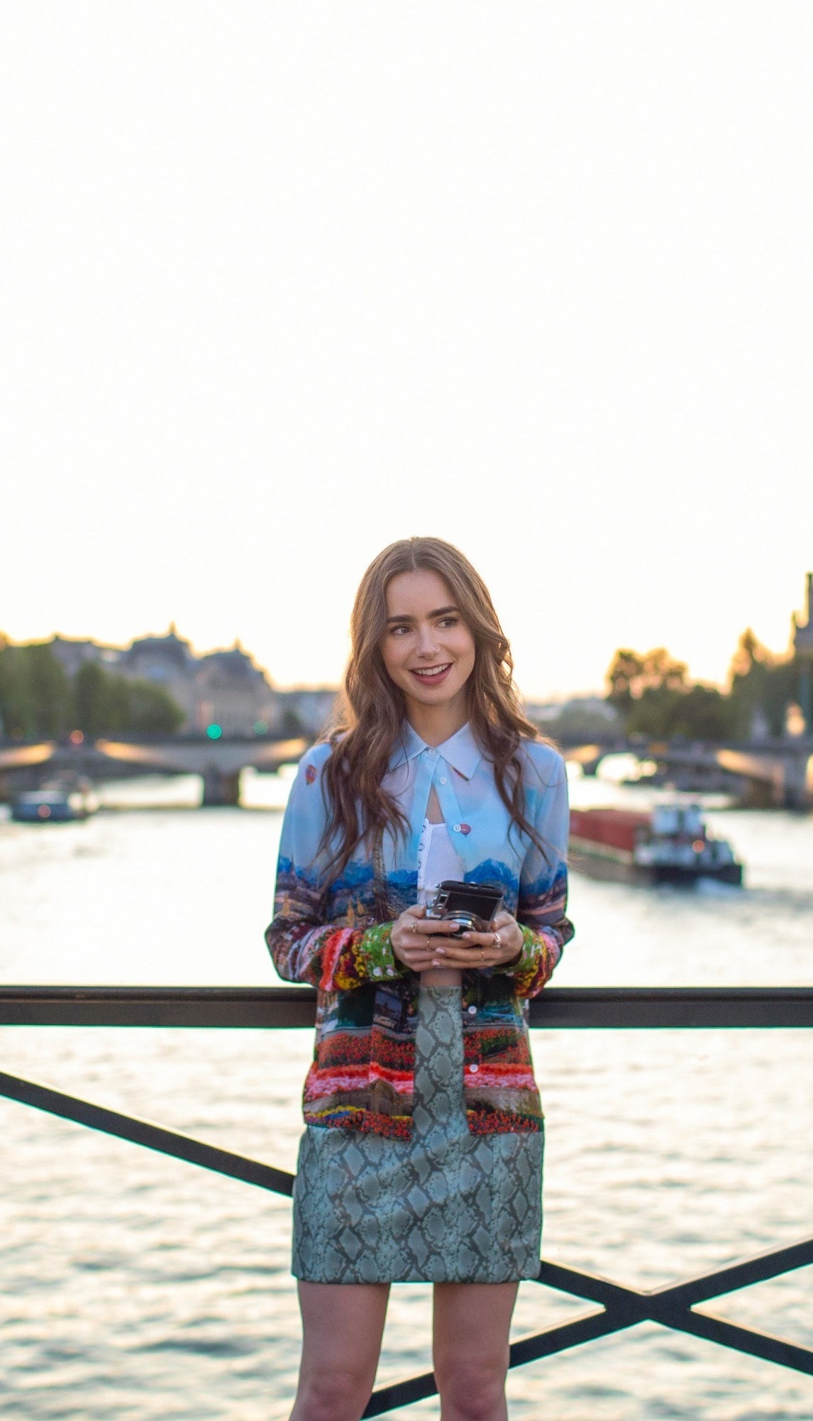 Emily stands on a bridge in front of the Seine wearing a snake print mini skirt and long sleeved Paris-motifed button up left open