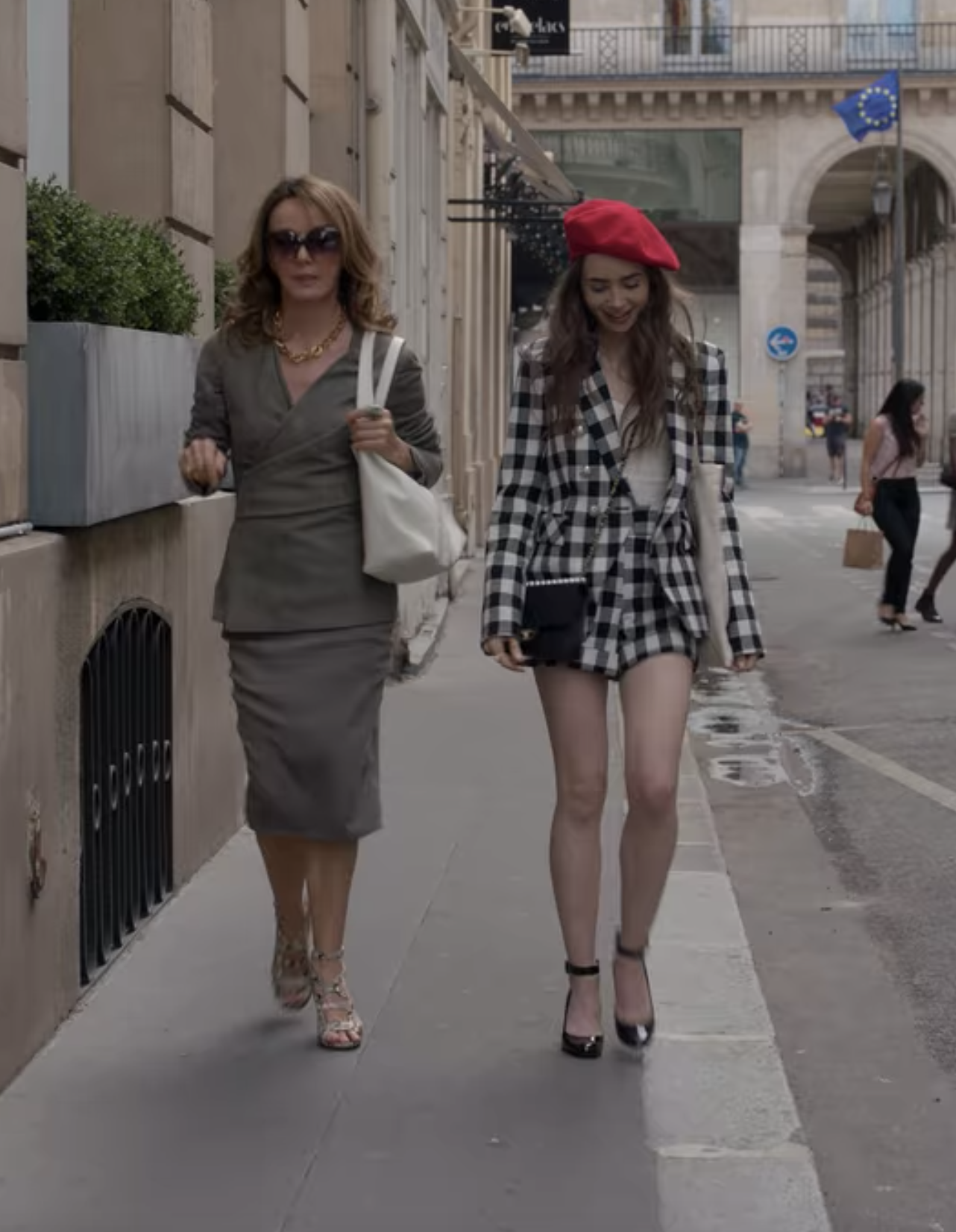 Emily and Sylvie walk down a street; Emily is dressed in a matching black and white gingham blazer/short set with a red beret and black heels