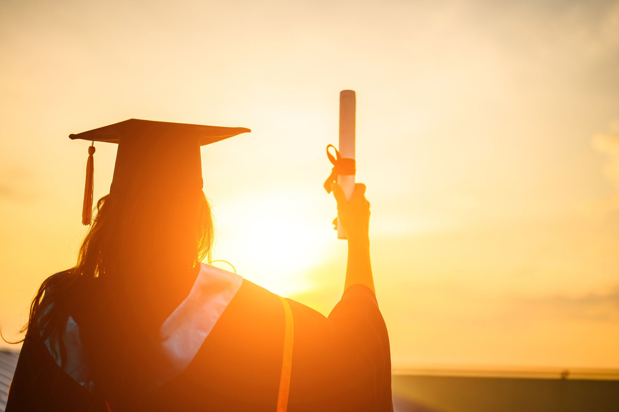 A person in a graduation cap and going holding a diploma in a sunset