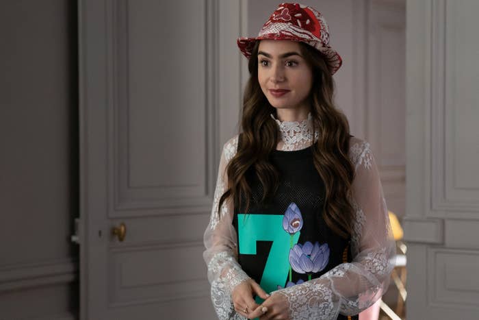 Emily wears a red paisley fedora and a black tank dress featuring a teal number 7 and flowers over a high necked, long sleeved sheer lace dress
