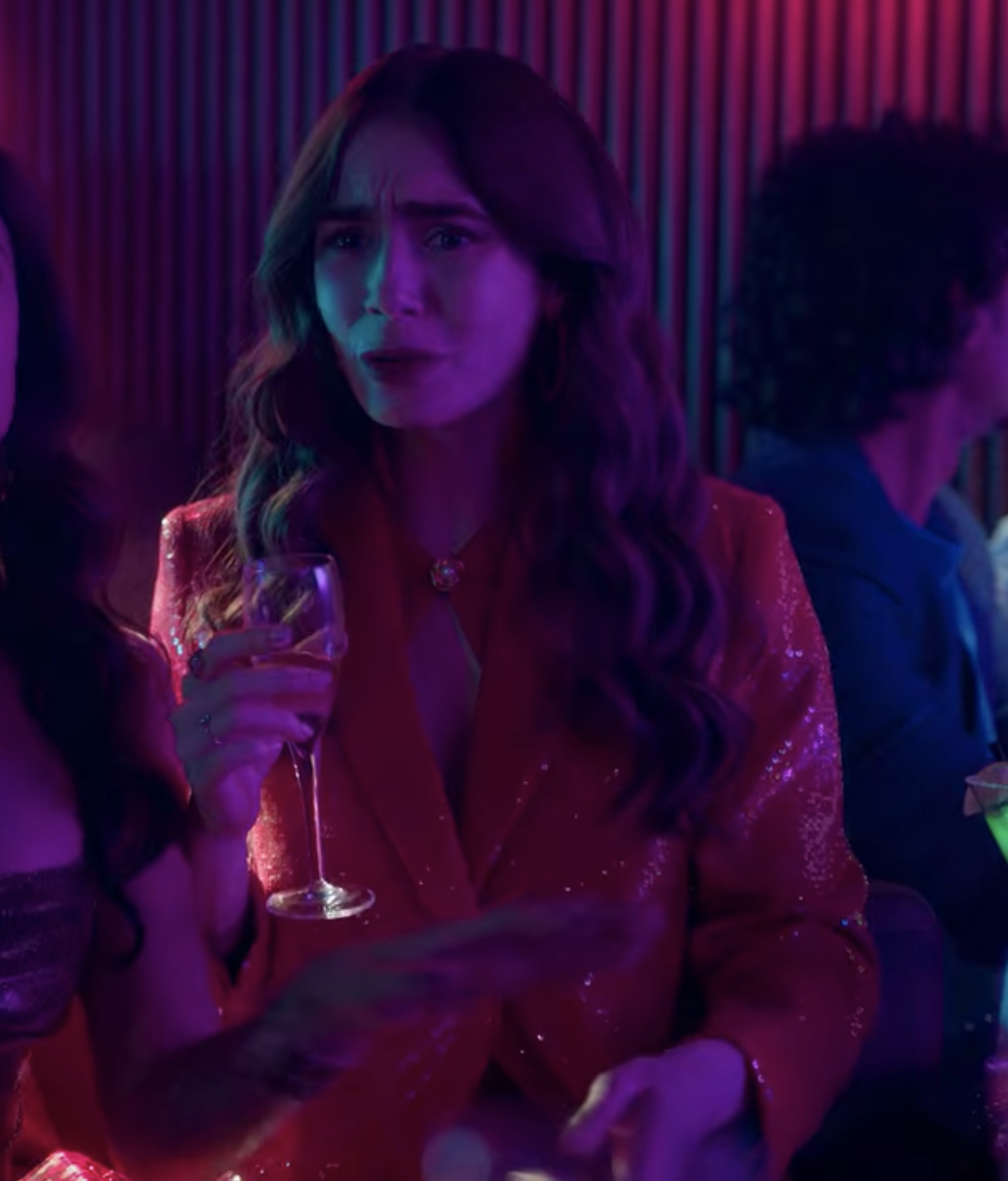 Emily holds a glass of champagne in a red, sparkly blazer with a red collar necklace at her throat