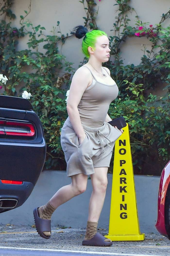 Billie Eilish, with neon-green hair tied up in a bun, baggy shorts, and socks and sandals, steps outside for a walk