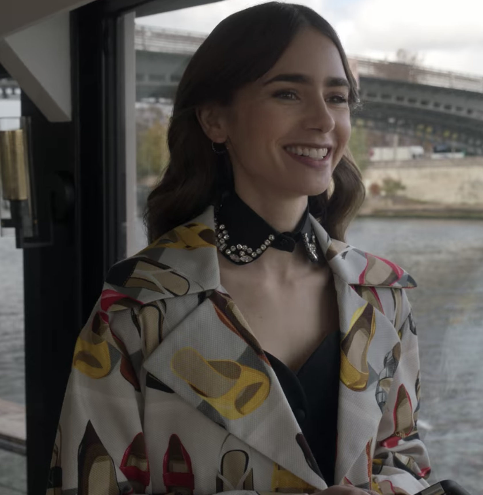 Emily smiles in front of the Seine wearing a beige coat with images of shoes on it and a black and rhinestone collared necklace