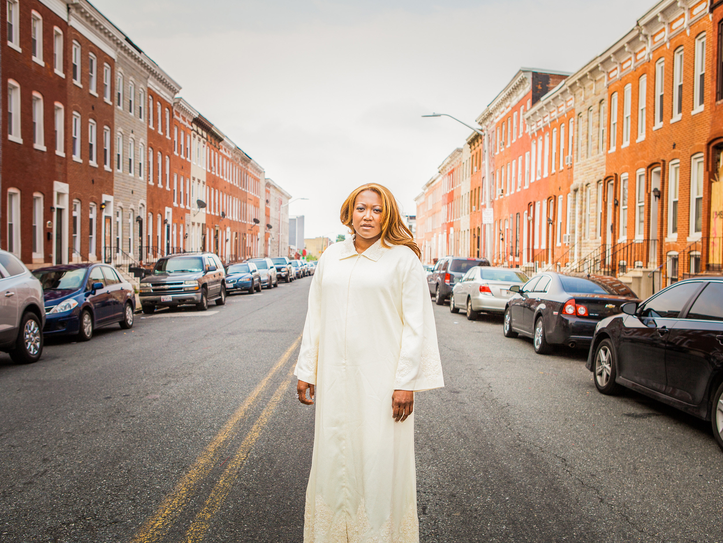 A woman in white robes on a street in Baltimore with brick row houses