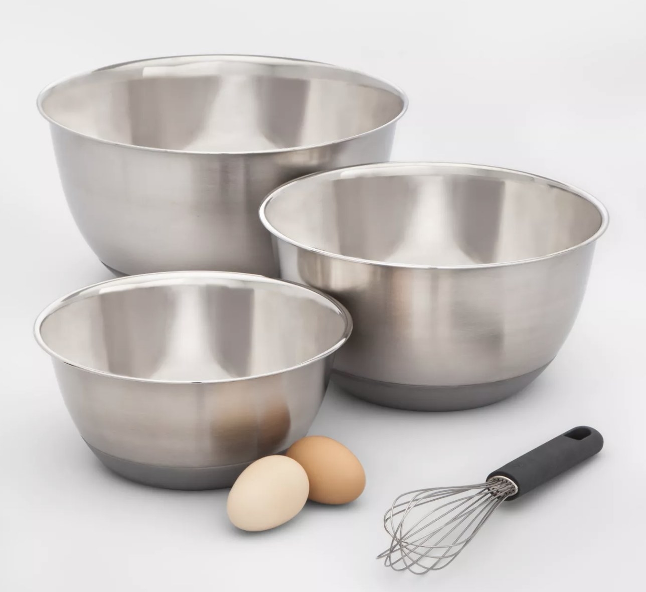 three different sized stainless steel bowls with grey silicone bases next to a whisk and eggs