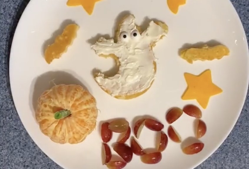 A plate of food including a clementine, a pancake shaped like a ghost and grapes that spell &quot;BOO&quot;