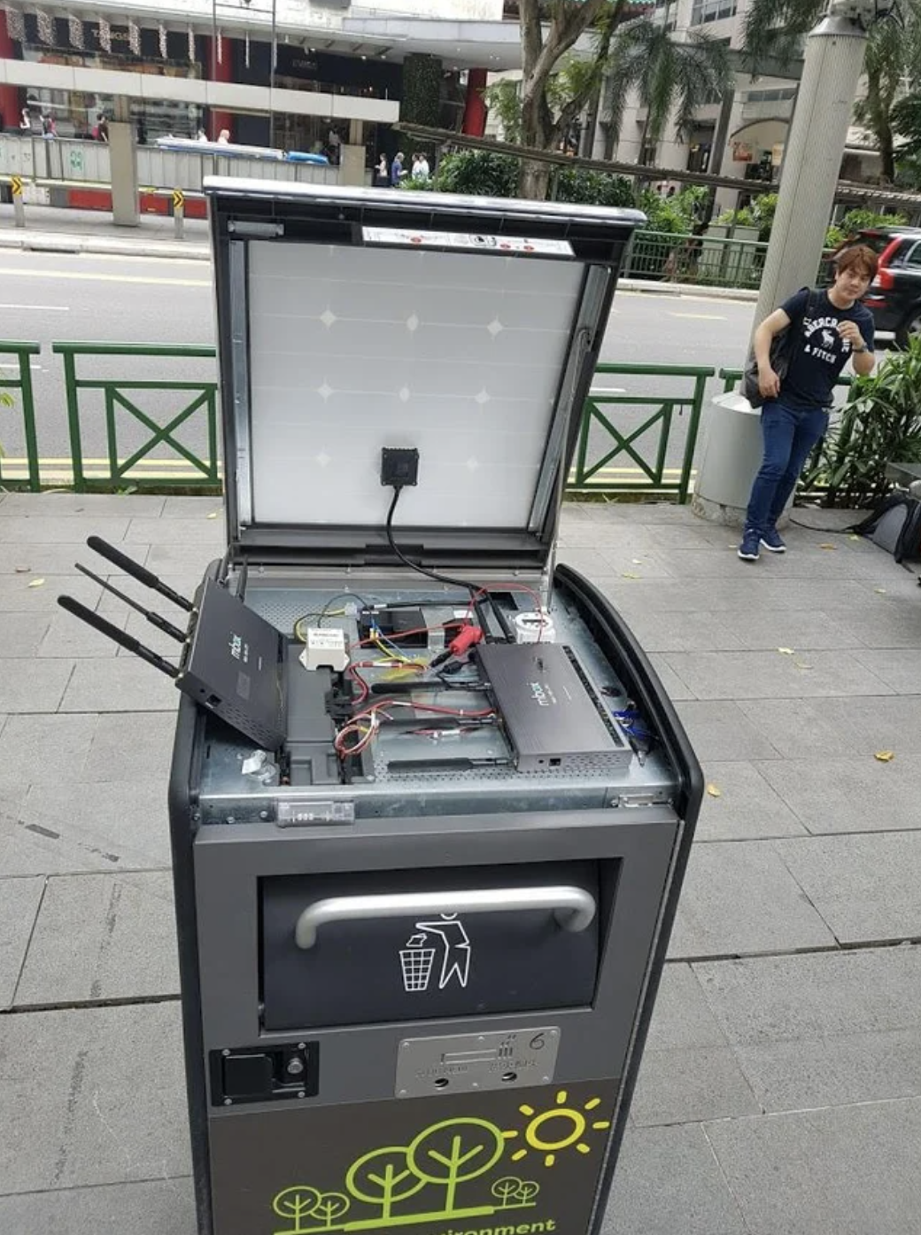 A public trash bin with modems, routers, and cords set up on top 