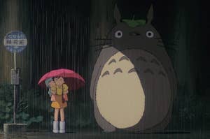 Satsuki, Mei, and Totoro wait at the bus stop in the rain