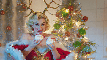 Katy Perry wearing reindeer antlers excitedly holding up two mugs of hot cocoa
