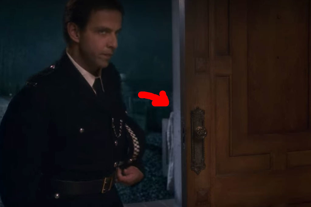 A police officer stands in the front doorway of Bly Manor; behind him a red arrow points to a portion of the little boy ghost that can be seen