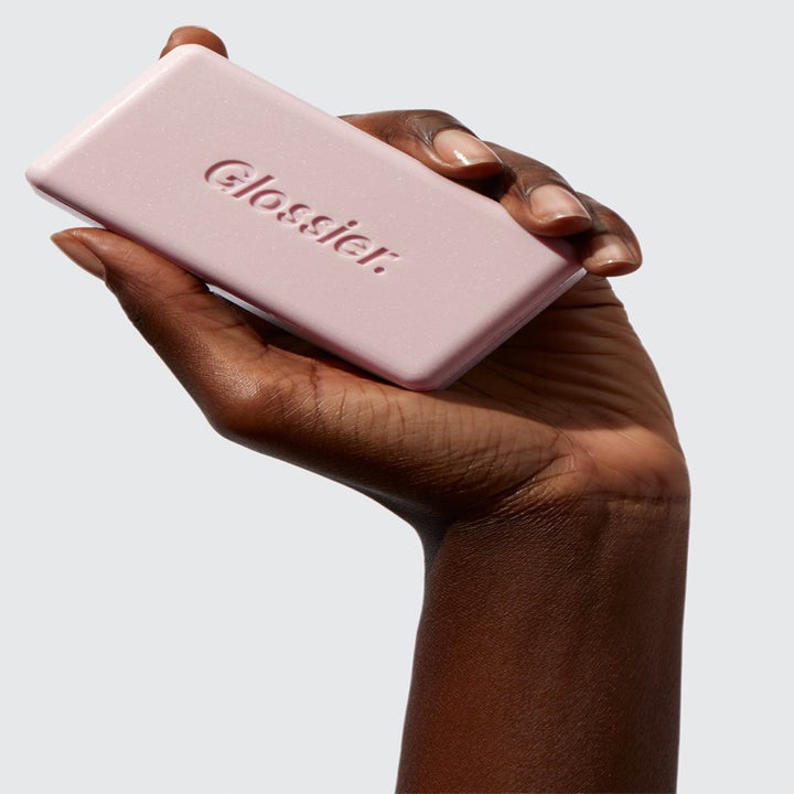 A hand holding the pink bar engraved with the Glossier logo