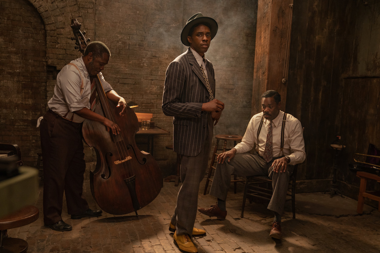 Chadwick Boseman wearing a suit and staring at the camera while men tune instruments behind him 