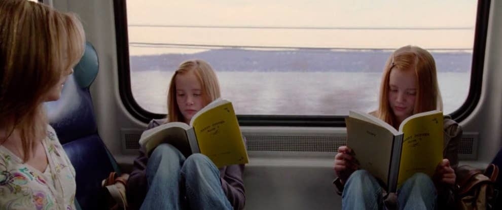 A set of twins are sitting on a train with their backs against the window while reading a book