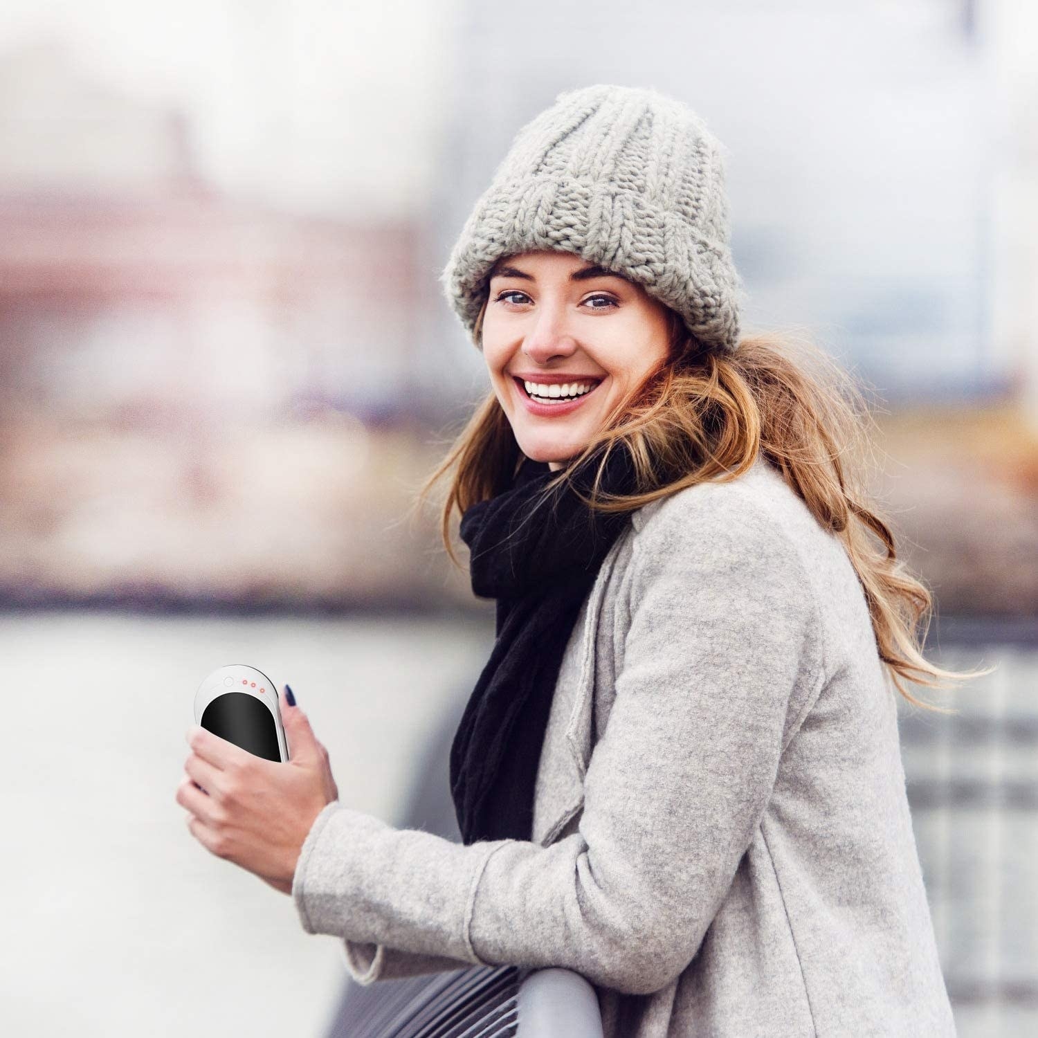 model smiling holding the infray hand warmer in their left hand while outside on a chilly day 