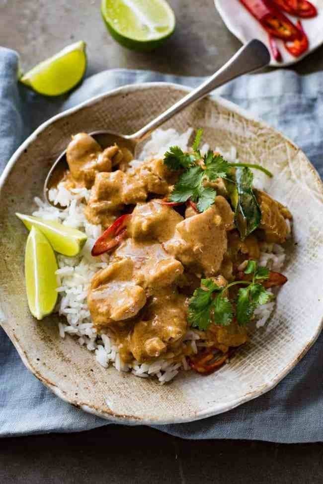 A bowl of rice topped with chicken curry, Thai chili pepper, fresh herbs, and lime wedges on the side.