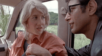 Laura Dern sitting in a car with Jeff Goldblum indicating a swooping motion over her head and laughing
