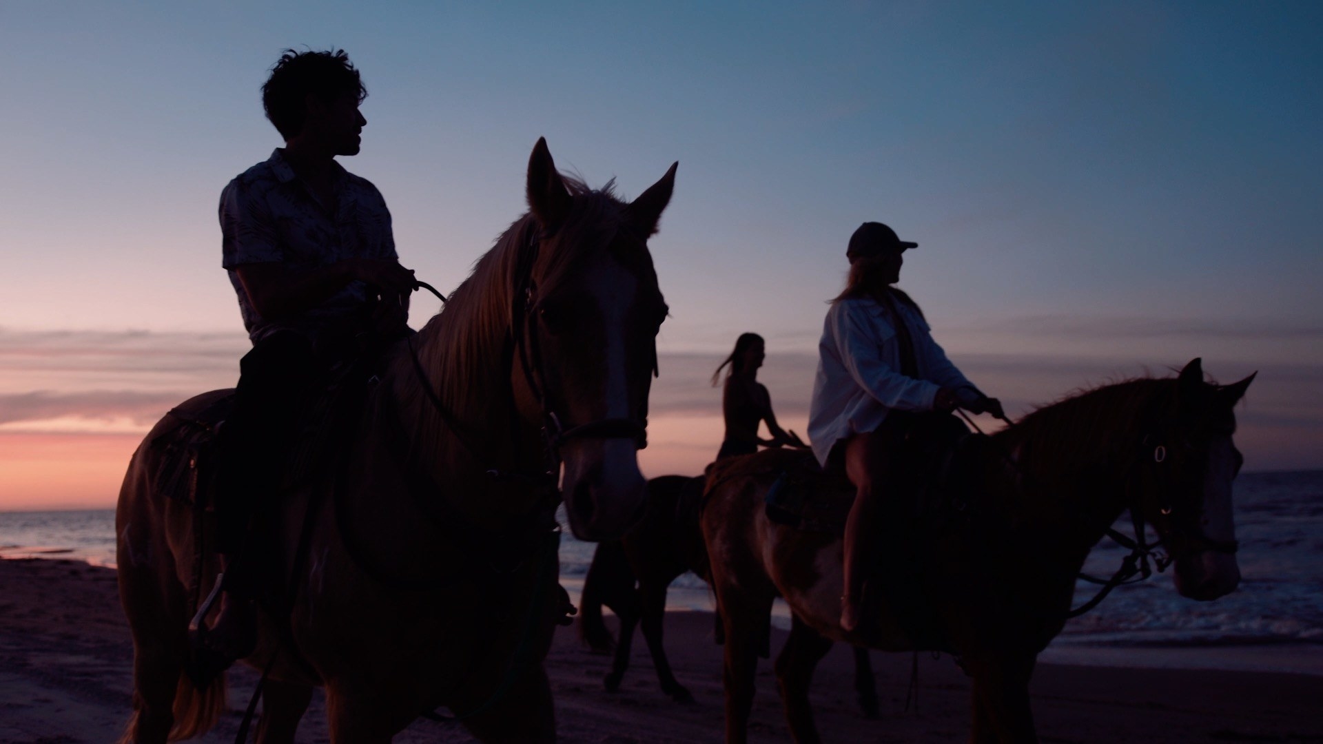Three young people riding horses by the beach at sunset