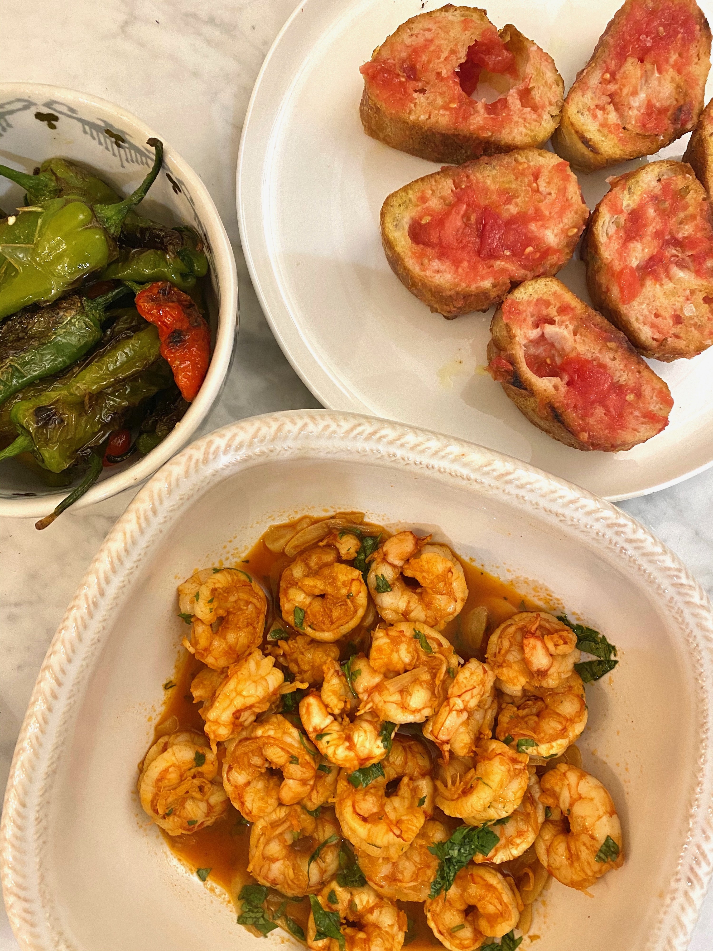 A plate of tomato bread, a bowl of shrimp in garlic sauce, and a third bowl of shishito peppers.