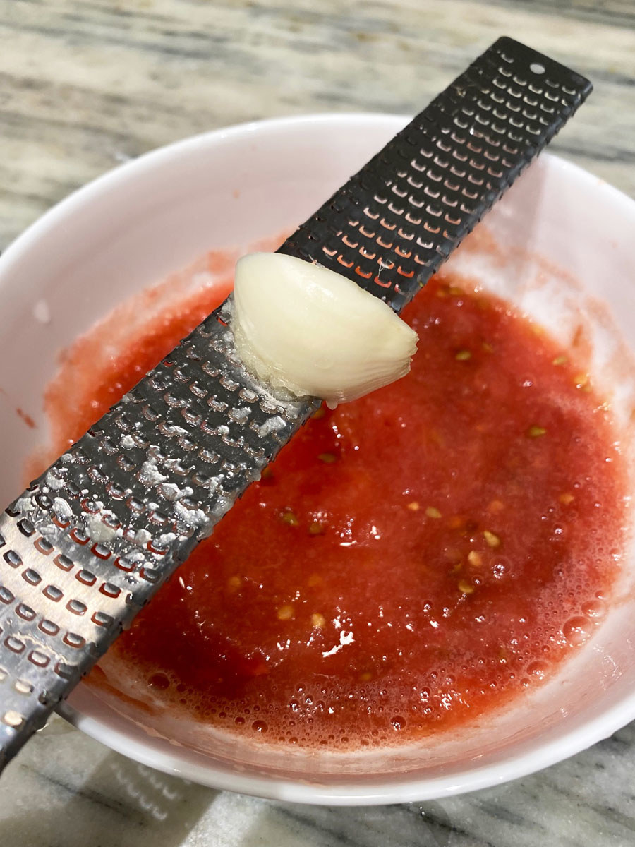 A clove of garlic being grated on a microplane into a bowl of tomato sauce.