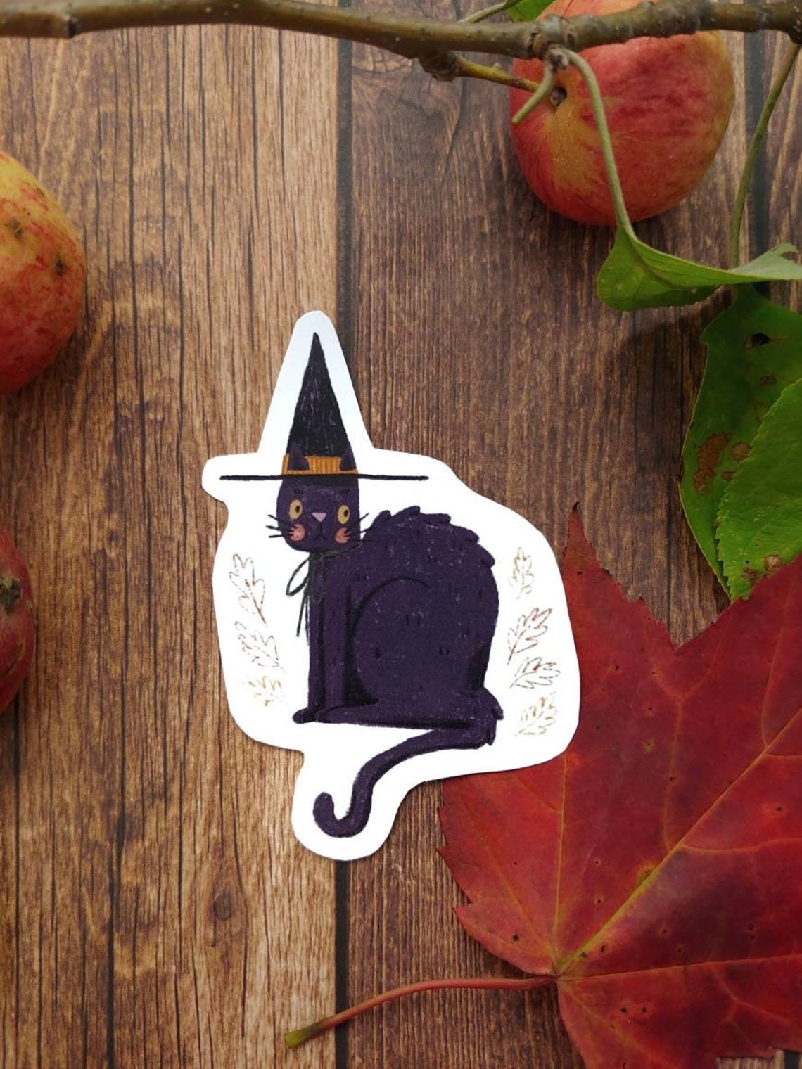 Picture of a deep purple cat on a white background with leaves, wearing a black witch&#x27;s hat.