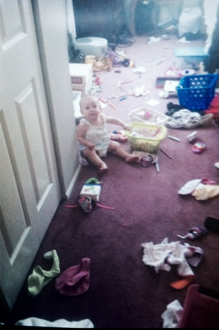 Foto antiga de um bebê em um cômodo com vários objetos jogados no chão.
