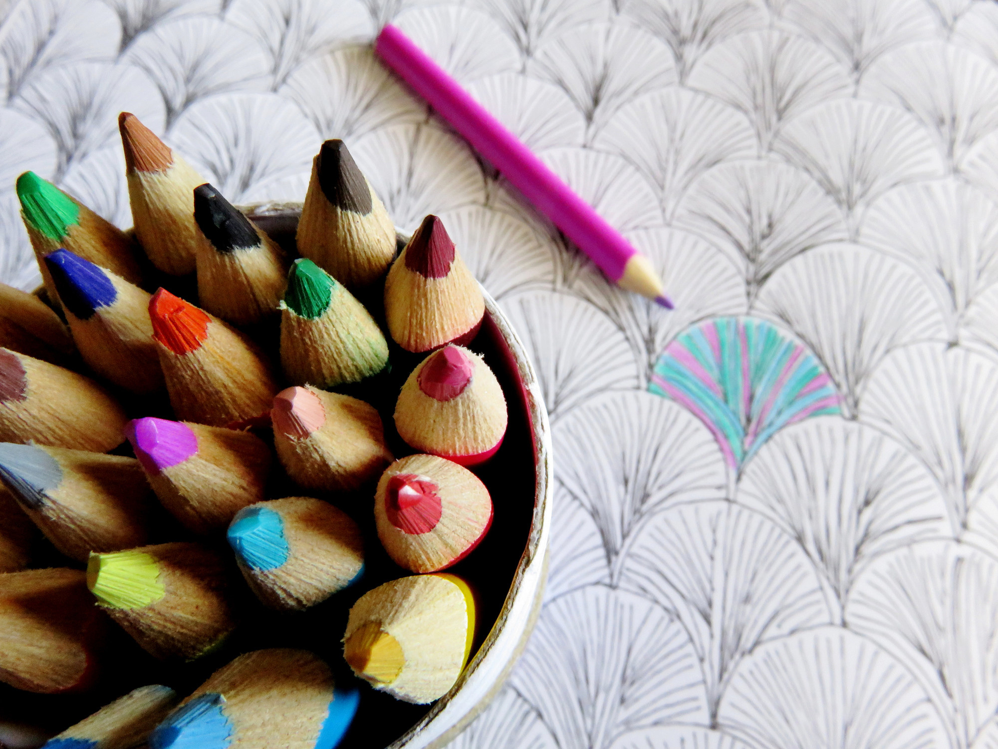 photo of coloring pencils in a can on the left corner while the can is on top of a table to draw on, in the middle is a pink pencil that has already colored in one portion of the table