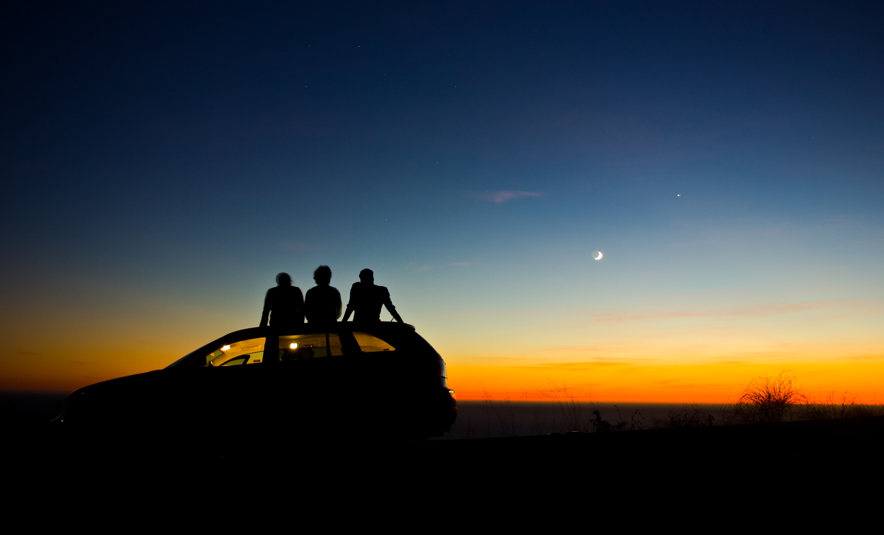 photo of the sunsetting and the moon appearing while on the left is a car with three people sitting on top of the roof of the car while watching the sunset, the sky is dark blue, blue, orange