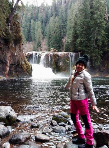 Reviewer wears same style pants in a hot pink shade with a white puffer jacket while hiking near a waterfall 