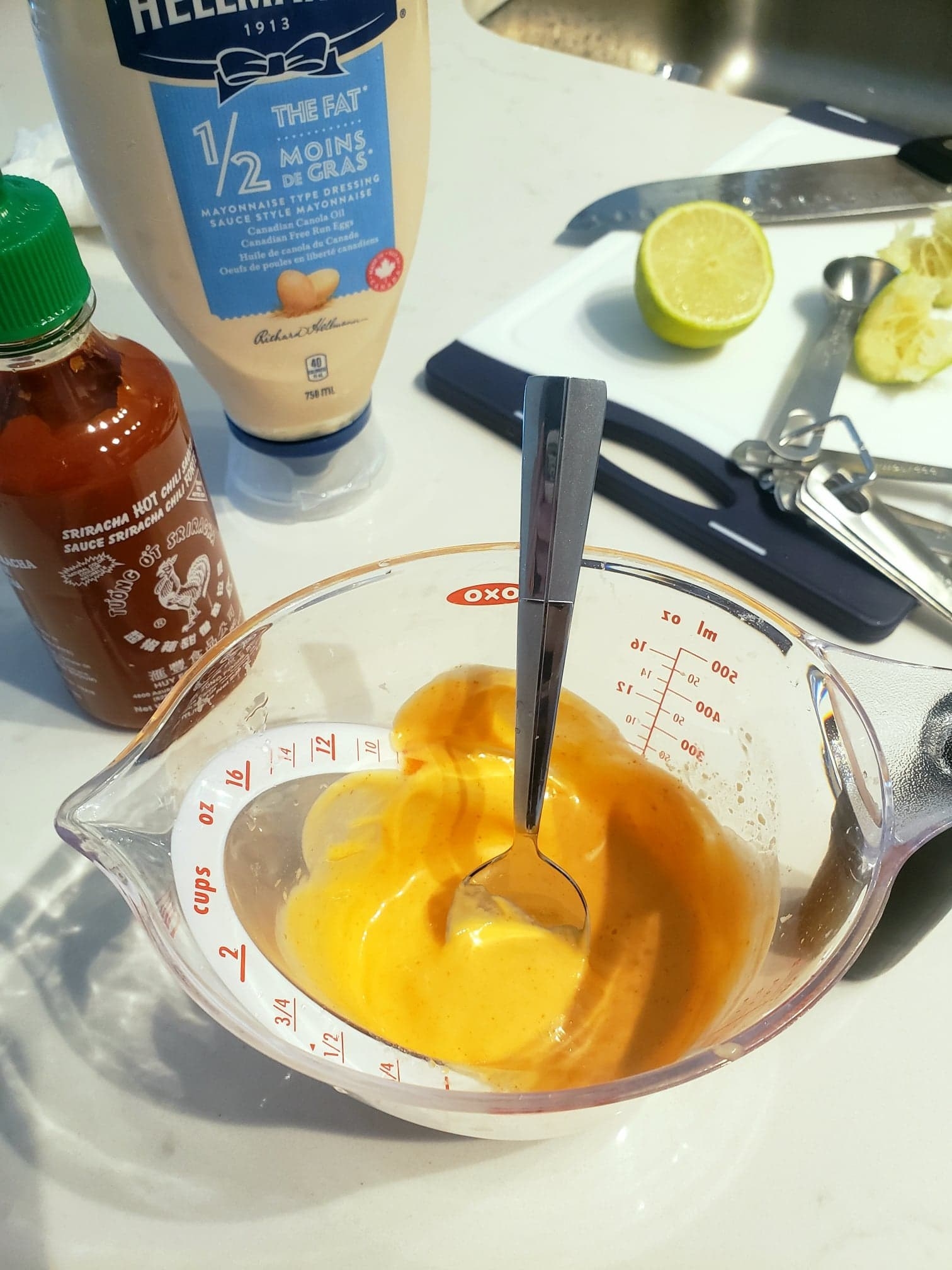 A measuring cup with Sriracha mayo freshly mixed inside, with the Sriracha bottle, mayonnaise bottle, and cut lime in the background