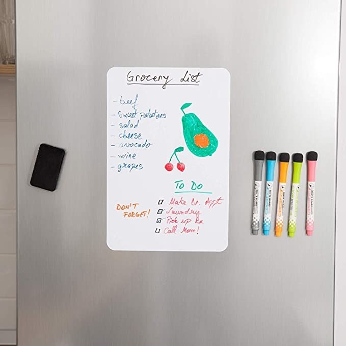 White board with a grocery list on it stuck to the fridge.