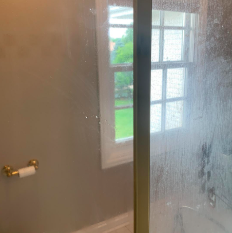 reviewer photo showing their glass shower door completely clear on the left side after using a Magic Eraser, and covered in build up on the right side where they have yet to use the Magic Eraser 