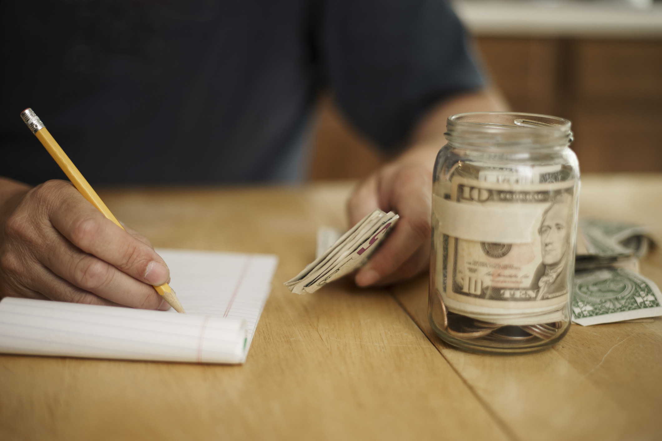 A person holding money and writing on a pad. A jar with cash inside