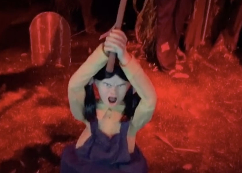 A creepy doll holds an axe in front of a graveyard scene
