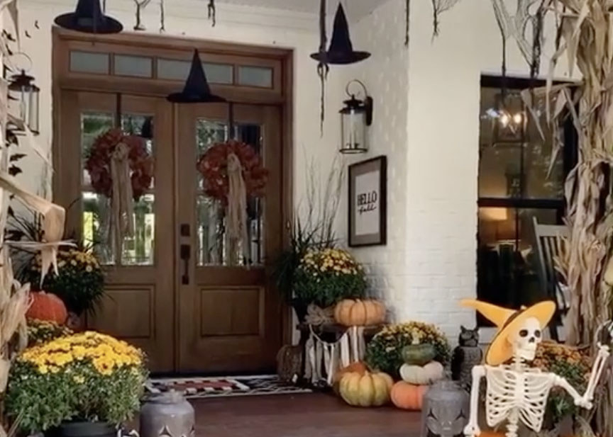 A stunning home with a porch full of yellow and red plants, pumpkins and a skeleton