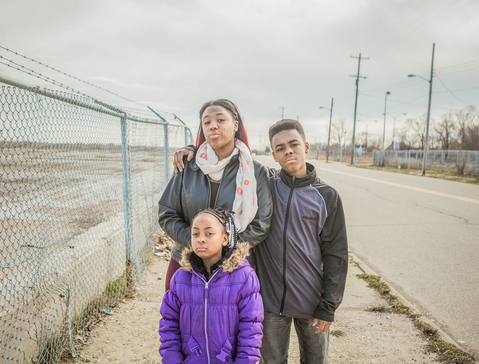 Three young people in coats on a sidewalk in Flint Michigan with a barbed wire fence